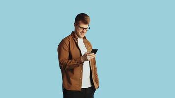 Young handsome caucasian man in glasses and casual clothes typing a message to a friend on his smartphone, looking at the camera and smiling on a blue background in the studio video