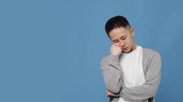 Young asian guy in casual clothes falls asleep standing, resting his head on his hand and then suddenly wakes up with a tired face on a blue background in the studio video