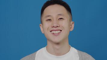 Portrait of a young asian smiling man close up standing and looking at the camera with a happy positive face on a blue background in the studio video