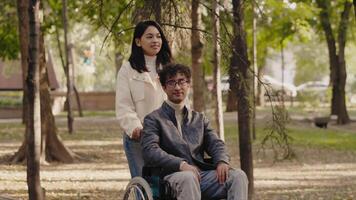 Young woman pushing a disabled young man in a wheelchair during a walk in the city park, and they enjoy spending time together. Concept of acceptance, love and support for people with disabilities video