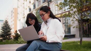 Young asian woman with a laptop sitting on a bench outdoors on a city street and explaining key points about work to her co-worker. Colleagues discussing working issues video
