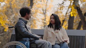 Young asian woman sitting on a bench and talking with a disabled young man in a wheelchair during a walk in the city park and enjoying spending time together video