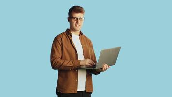 Young attractive man in glasses and casual clothes typing on a laptop which holding in his hands, looking at the camera and extending his hand for a handshake with a smile on a blue background video