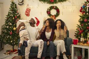 A lively Christmas celebration with three african friends enjoying drinks and holiday cheer on a sofa. The festive atmosphere includes Christmas trees, wreaths, lights, and decorations. photo
