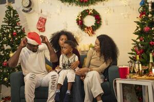 A lively Christmas celebration with three african friends enjoying drinks and holiday cheer on a sofa. The festive atmosphere includes Christmas trees, wreaths, lights, and decorations. photo
