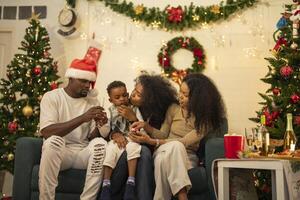 A lively Christmas celebration with three african friends enjoying drinks and holiday cheer on a sofa. The festive atmosphere includes Christmas trees, wreaths, lights, and decorations. photo
