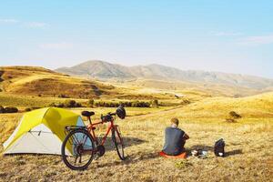 Adventurous cyclist sit by touring bicycle in mountains on side of green tent have break time rest in morning. Solo travel journey. Long-term travel around the world photo