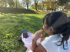 Asian little girl on the grass using phone in outdoor making call with love sign hand gesture photo