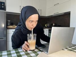 Asian Muslim woman in hijab drinking coffee and using laptop at home kitchen with serious face looking at the laptop screen. Working mom from home. photo