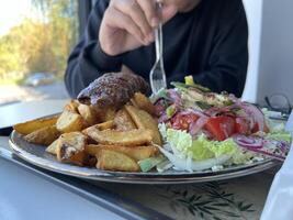 Unrecognized man holding plastic fork for enjoying Kebab with French fries and salad on a plate on white table in a restaurant photo