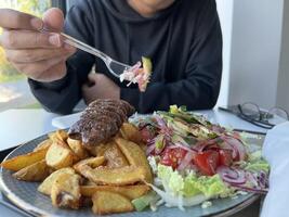 Unrecognized man holding plastic fork. With served Kebab with French fries and salad on a plate on white table in a restaurant photo