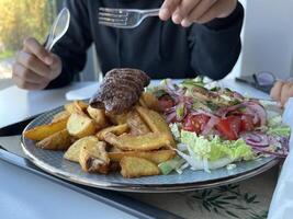 Unrecognized man holding plastic fork. With served Kebab with French fries and salad on a plate on white table in a restaurant photo