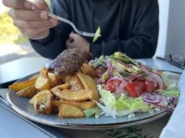 Unrecognized man holding plastic fork. With served Kebab with French fries and salad on a plate on white table in a restaurant photo