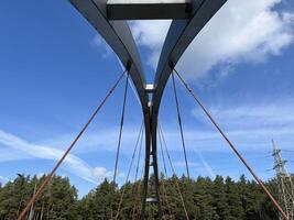 A close-up view of a modern bridge with a distinctive arch design with a clear blue sky with clouds and a line of trees in the distance. A high-voltage power line is also visible in the background photo