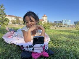 Smiling Asian little girl on the grass using phone in outdoor making call with love sign hand gesture photo