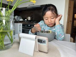 Asian little girl watching mobile phone while eating in the modern kitchen. Tech-related health issues concept. photo