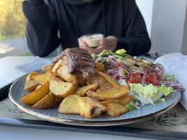 Unrecognized man holding kebab sauce for enjoying Kebab with French fries and salad on a plate on white table in a restaurant photo