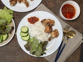 Nasi ayam and lalaban include a plate of rice with fried chicken thigh sprinkled with onions and parsley on a white plate served with tomato sauce, cucumber and lettuce. photo