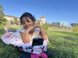 Smiling Asian little girl on the grass using phone in outdoor making call with love sign hand gesture photo
