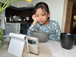 Asian little girl watching mobile phone after eating in the modern kitchen. Screen time health risks concept photo