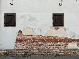 A weathered wall with peeling white paint, exposing red brick. Two small windows with dark shutters and vegetation at the base ideal for creative projects like interior design or backgrounds photo