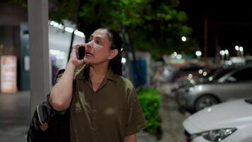 A woman waves her hand to signal her pickup location at a gas station while talking on the phone at night. Capturing an urban moment of waiting, communication, and night scene. video