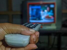 A hand holding a worn-out TV remote control, pointing it towards a blurry television screen, capturing a nostalgic moment of traditional TV watching. photo