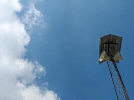 A tall communication tower with dual loudspeakers mounted under a metal roof, set against a bright blue sky with light clouds. The contrast between the man-made structure and natural sky. photo