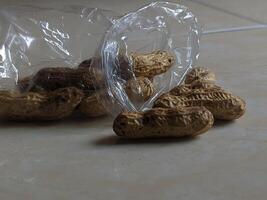 A close-up of raw peanuts in a transparent plastic bag, scattered on a light-colored surface. The peanuts are in their shells and have a natural, earthy tone. photo