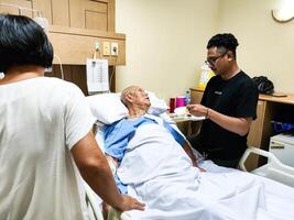 Caregiver Assisting Elderly Patient with Meal in Hospital Room with a Visitor photo