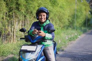 Online taxi rider wearing green jacket and helmet, riding motorcycle, using a mobile phone while on the way to pick up customer. Transportation application concept. photo