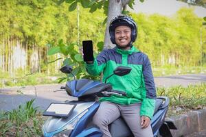 Online taxi rider wearing green jacket and helmet, riding motorcycle, showing blank screen mobile phone while on the way to pick up customer. Transportation application concept. Smartphone mockup photo