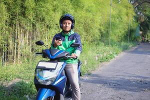 Online taxi rider wearing green jacket and helmet, riding motorcycle, using a mobile phone while on the way to pick up customer. Transportation application concept. photo