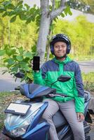 Online taxi rider wearing green jacket and helmet, riding motorcycle, showing blank screen mobile phone while on the way to pick up customer. Transportation application concept. Smartphone mockup photo