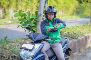 Online taxi rider wearing green jacket and helmet, riding motorcycle, checking time on his watch while on the way to pick up customer. Transportation application concept. photo