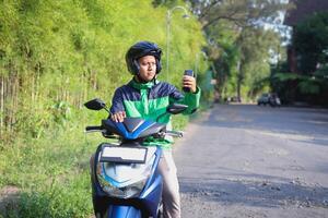 Online taxi rider wearing green jacket and helmet, riding motorcycle, using a mobile phone while on the way to pick up customer. Transportation application concept. photo