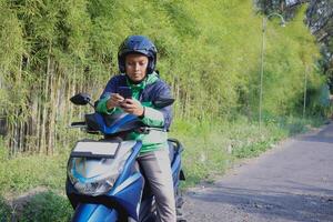 Online taxi rider wearing green jacket and helmet, riding motorcycle, using a mobile phone while on the way to pick up customer. Transportation application concept. photo