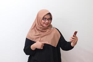 Portrait of cheerful Asian woman with hijab standing against white background, pointing away with her finger while using mobile phone photo