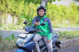 Online taxi rider wearing green jacket and helmet, riding motorcycle, using a mobile phone while on the way to pick up customer. Transportation application concept. photo