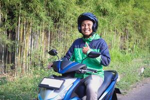 Online taxi rider wearing green jacket and helmet, riding motorcycle while on the way to pick up customer and showing thumb up hand gesture. Transportation application concept. photo