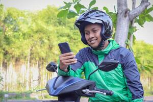 Online taxi rider wearing green jacket and helmet, riding motorcycle, using a mobile phone while on the way to pick up customer. Transportation application concept. photo