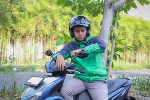 Online taxi rider wearing green jacket and helmet, riding motorcycle, checking time on his watch while on the way to pick up customer. Transportation application concept. photo