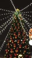 View from the bottom on the beautiful Christmas tree with colorful decorations and garland of white lights standing on the square. Concept. Beautiful snowy winter scenery of Christmas holiday video