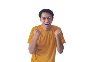 Handsome Asian man in a orange shirt raises both fists in a celebratory gesture, his face beaming with excitement and on white background emphasizes his triumphant and enthusiastic expression. photo