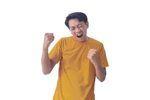 Handsome Asian man in a orange shirt raises both fists in a celebratory gesture, his face beaming with excitement and on white background emphasizes his triumphant and enthusiastic expression. photo