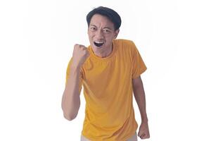 Handsome Asian man in a orange shirt raises both fists in a celebratory gesture, his face beaming with excitement and on white background emphasizes his triumphant and enthusiastic expression. photo
