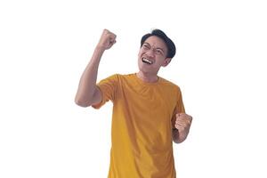 Handsome Asian man in a orange shirt raises both fists in a celebratory gesture, his face beaming with excitement and on white background emphasizes his triumphant and enthusiastic expression. photo