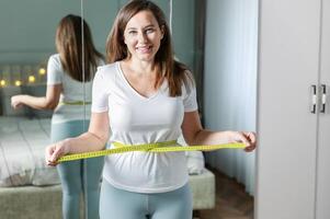 Smiling young woman after weight loss measuring waist in front of mirror photo