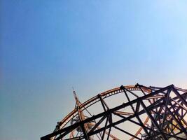 A roller coaster is seen in the distance with a clear blue sky in the background. The roller coaster is a large structure with a lot of metal and it is surrounded by a lot of empty space photo