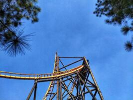 A roller coaster is seen from below with a blue sky in the background. The roller coaster is yellow and has a lot of metal photo